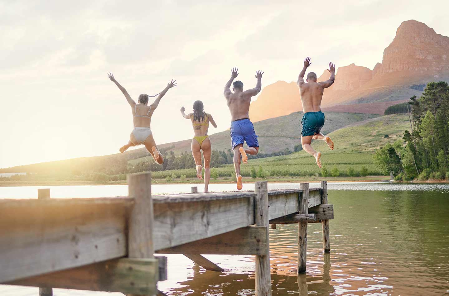 Vier Freunde springen von einem Steg in den See. Im Hintergrund sind Berge zu sehen, die von der untergehenden Sonne angestrahlt werden.