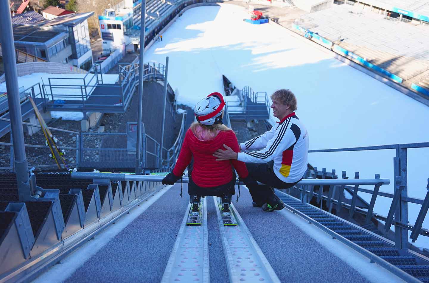 Peter Tümmers auf der Skisprungschanze in Oberstdorf. Von hinten ist eine Frau im roten Parka zu sehen, die sich bereit macht, die Schanze hinunter zu fahren.