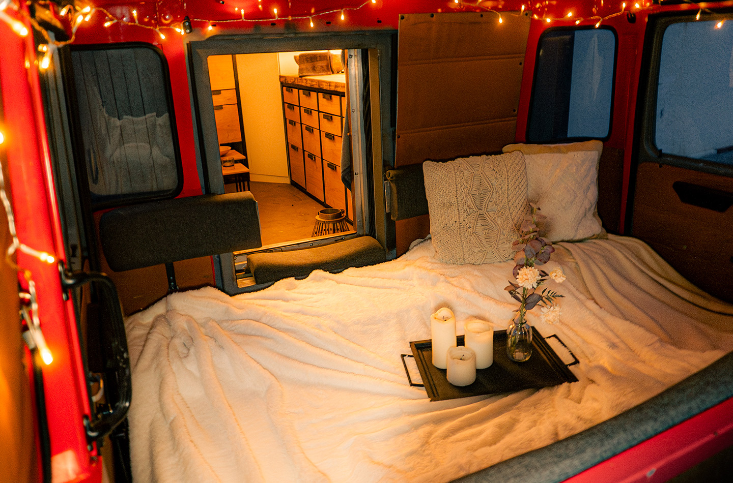 Beleuchtetes Bett mit Kissen, Tablett mit Kerzen und Blumen. Im Hintergrund Kommode.