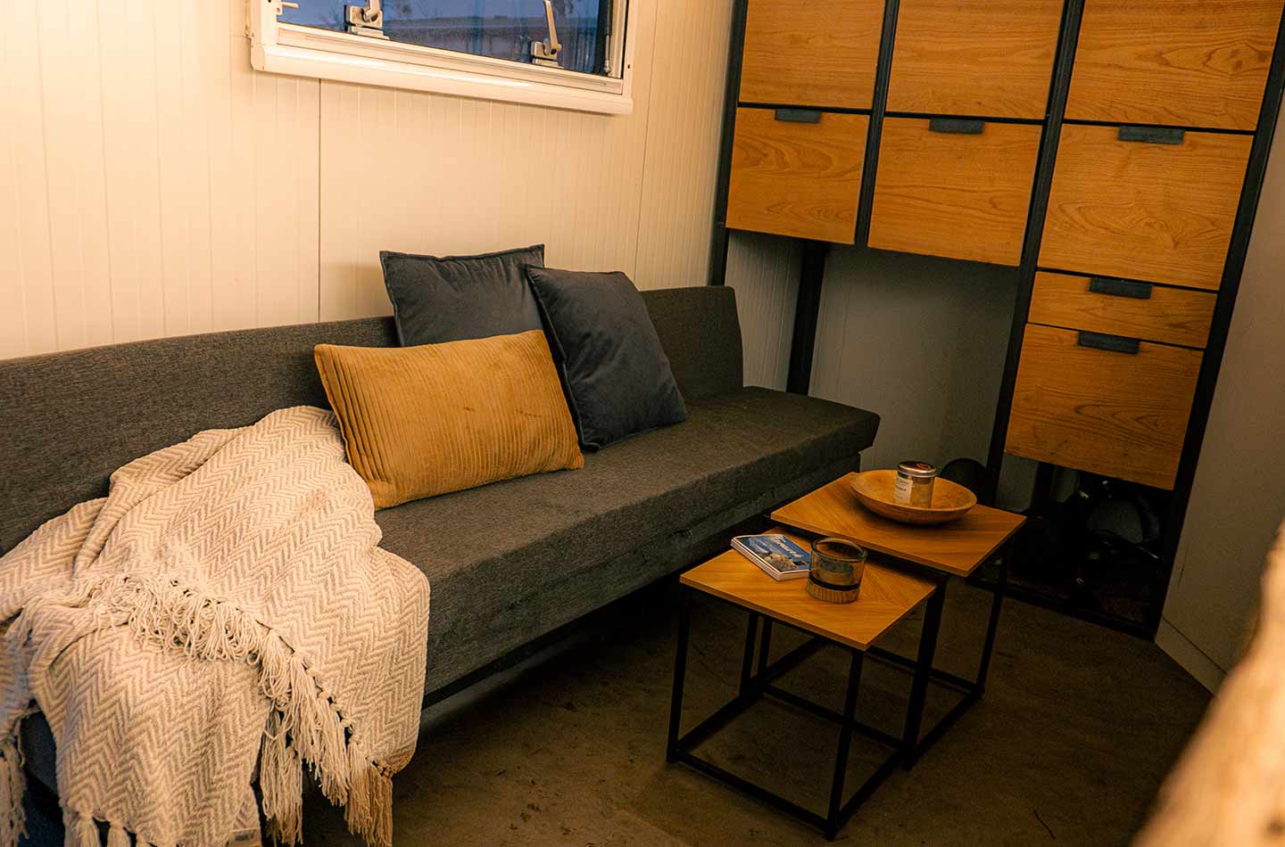 Couch mit Kissen und Decke. Kleine Tische und im Hintergrund ein Schrank mit Holzverkleidung.