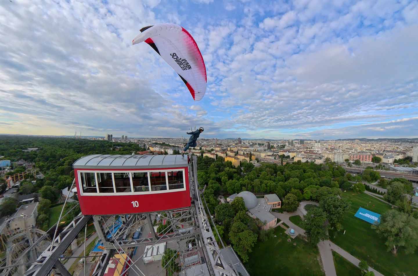 Aussicht über Wien. Riesenrad mit rot-weißem Wagon. Gleitschirmflieger springt ab. 