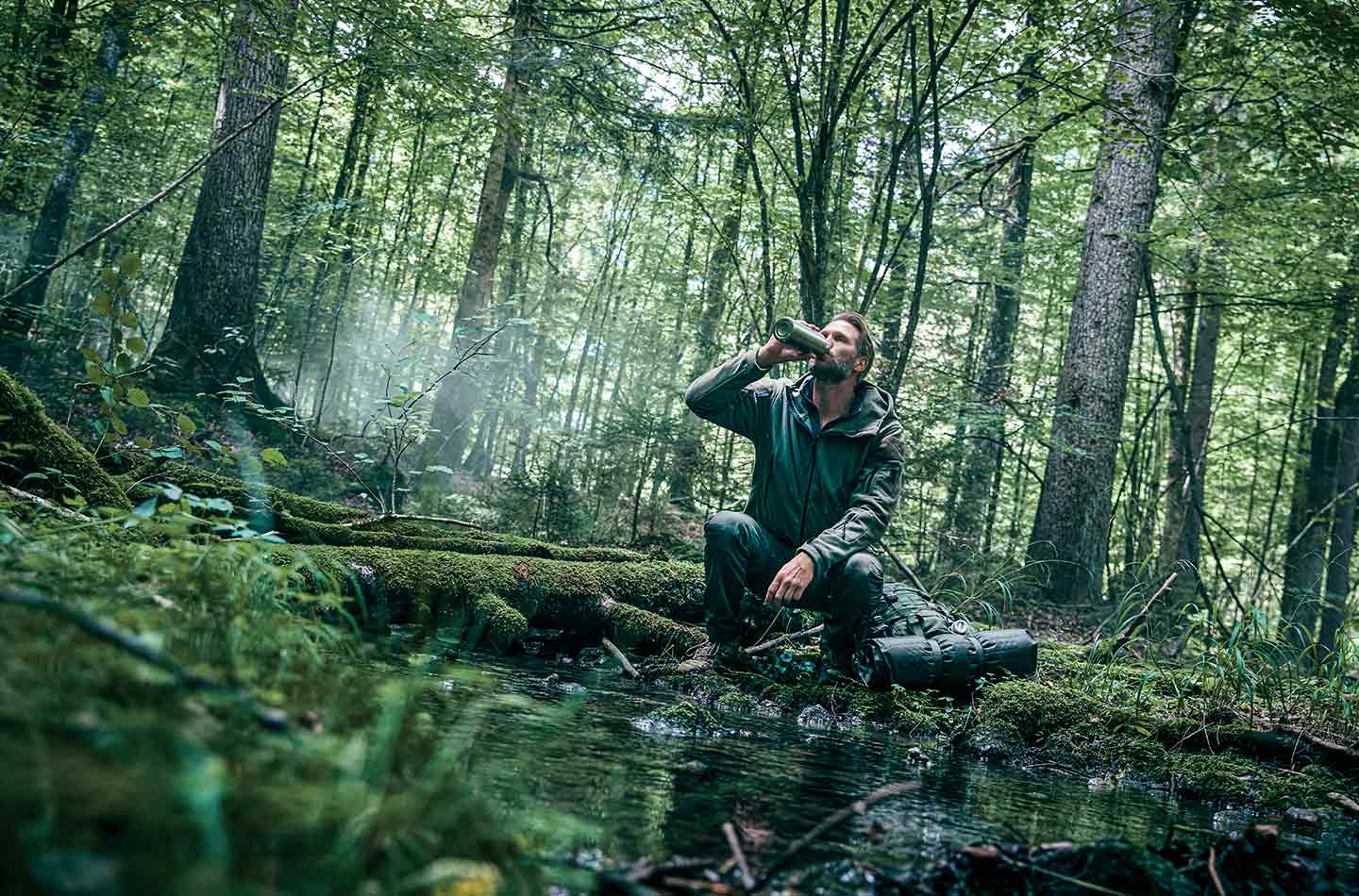 Ein Mann trinkt Wasser an einem Bach im Wald